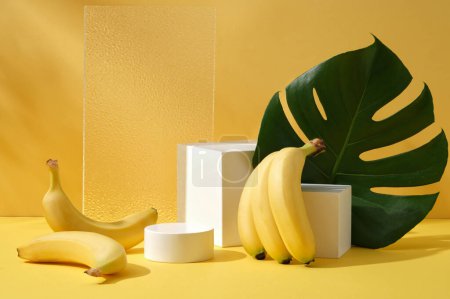 Foto de Fondo amarillo para la publicidad de productos cosméticos de ingrediente de plátano con podios blancos, lámina acrílica y hojas de la selva monstera. Espacio para el diseño - Imagen libre de derechos