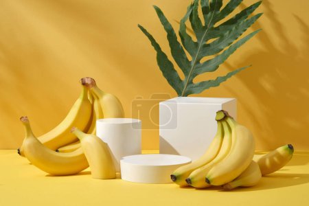Photo for Bunch of bright yellow tiny bananas decorated on a yellow background with cylinder white podiums and green leaf. Minimal scene for cosmetic product presentation. Front view - Royalty Free Image