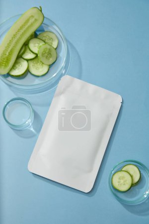 Mockup of a cucumber sheet mask on blue background with cucumber slices in petri dishes. Templates for beauty product packaging based on cucumber, natural extract good for skin and hair.