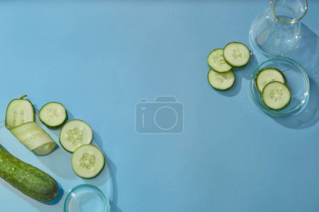 Téléchargez les photos : Vue de dessus des tranches de concombre décorées avec de la verrerie de laboratoire sur fond bleu. Espace pour le produit d'affichage. La haute teneur en eau et les antioxydants du concombre rendent la peau élastique et flexible. - en image libre de droit