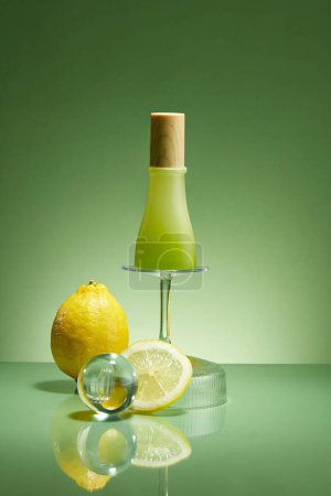 Green background for branding and minimal presentation of product from lemon ingredient. Glass bottle unlabeled on glass cup upside down decorated with glass ball and fresh lemon. Mockup, copy space