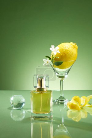 Foto de Escena para la publicidad de productos de aceite de fragancia de aceite de limón. Prototipo de botella de perfume sin etiquetar con taza de vidrio que contiene limón y flor blanca, bola de cristal y cáscara de limón sobre fondo verde - Imagen libre de derechos