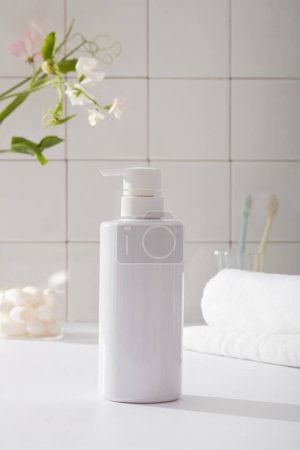 Front view of white bottle unlabeled in bathroom background with towel, toothbrush and flower branch on white tile wall. Mockup for cosmetic product with beauty concept.