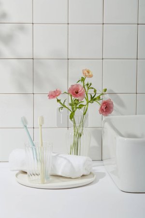 Photo for Beauty photo with bathroom concept. Toothbrushes in glass cup, white towel on round dishes, one corner of the washbasin and pink flower vase on tile wall background. Front view, copy space - Royalty Free Image