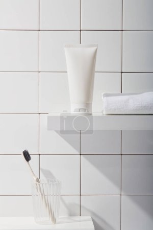 Photo for Front view of white plastic tube unlabeled and cotton towel on pedestal, under is toothbrush in glass cup on tile wall background. Mockup for cosmetic product with bathroom concept. - Royalty Free Image