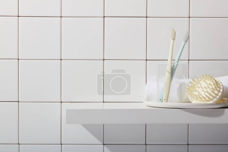Photo for Bathroom background with toothbrushes in glass cup, scalp massage brush and towel on white pedestal on tile wall background. Empty space for display cosmetic product. Front view, copy space. - Royalty Free Image