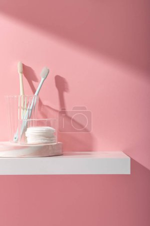 Téléchargez les photos : Scène publicitaire pour les soins buccodentaires et les produits de nettoyage avec des brosses à dents dans une tasse en verre et enlever le coton de maquillage sur piédestal, sur fond rose. Espace pour le texte et le design. - en image libre de droit