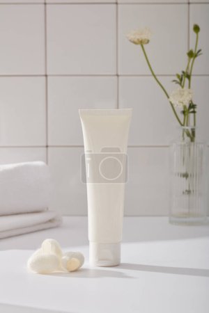 Photo for A white plastic tube unlabeled mockup for facial moisturizer cream or facial cleanser, towel, flower vase and silkworm cocoons decorated on white tile background. Bathroom concept, daily routine content - Royalty Free Image