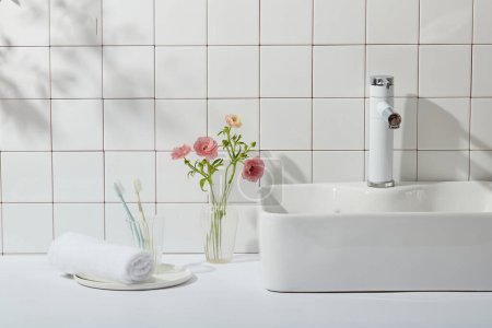 Téléchargez les photos : Intérieur d'une salle de bain moderne avec un mur de tuiles blanches, lavabo, serviette et brosses à dents. Vase en verre avec des fleurs roses qui se détachent sur fond blanc. Le concept de salle de bain, espace de copie - en image libre de droit
