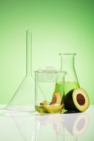Photo for Front view of lab glassware containing essential oil of avocado and fresh organic avocado decorated on green background. Researching natural essential oils to make cosmetics in the laboratory - Royalty Free Image