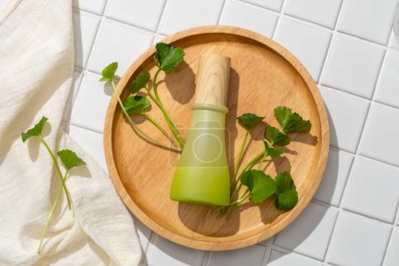 Concept de salle de bain pour la publicité cosmétique de l'extrait de gotu kola - bouteille en verre vert sur des plats en bois avec feuilles de gotu kola et serviette beige sur fond de carrelage blanc.