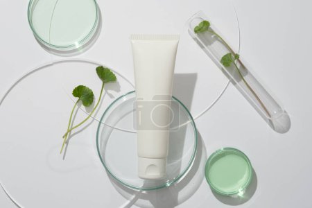 Photo for Cosmetic advertising scene with unlabeled white plastic tube placed on laboratory glassware and gotu kola leaves on white background. Copy space. Natural components for cosmetics - Royalty Free Image