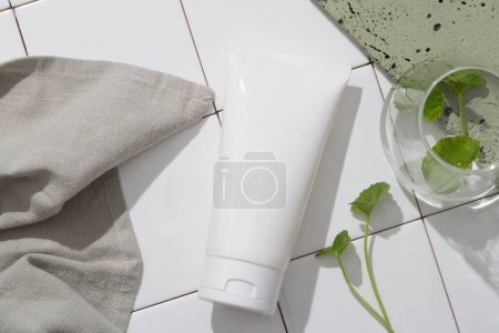 Photo for Bathroom concept for advertising cosmetic of gotu kola extract - plastic tube unlabeled, gray towel, gotu kola leaves on white tile floor. Cosmetic, beauty product promotion mockup. - Royalty Free Image