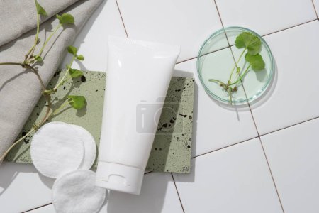 Photo for Bathroom concept with unlabeled plastic tube on gray brick, gotu kola leaves, towel and cotton pad on a white tile floor. Mockup for promoting cosmetic and beauty products with natural extract - Royalty Free Image