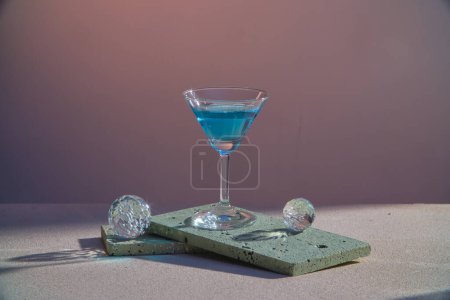 Téléchargez les photos : Boisson cocktail bleu dans un verre avec boule transparente placée sur des briques rectangle gris sur fond violet foncé. Photo publicitaire avec un concept minimal. Espace de copie. - en image libre de droit