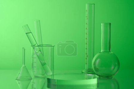 Science and medical background with lab glassware - test tubes, boiling flask, beaker and round transparent podium on green background. Blank space for product presentation.
