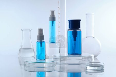 Mockup scene for cosmetic with three blue spray bottle without label placed on transparent podium and laboratory glassware filled colorless liquid decorated on white background. Front view, copy space