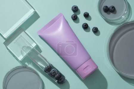 Mockup scene with purple bottle for cosmetics of blueberry extract on blue background. Petri dish filled essence and test tube decorated with fresh blueberries. Space for design