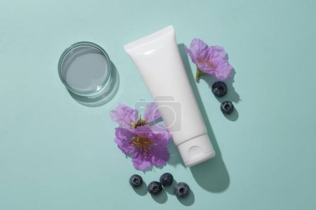 Minimal backdrop for cosmetic branding presentation - white plastic tube unlabeled container cleanser or cream, blueberries and purple flowers decorated on blue background.