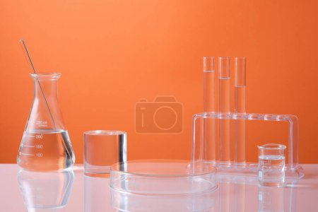 Photo for Front view of laboratory equipment - erlenmeyer flask, test tubes and beaker filled colorless liquid and cylinder podium for product presentation on orange background. Science and medical background - Royalty Free Image