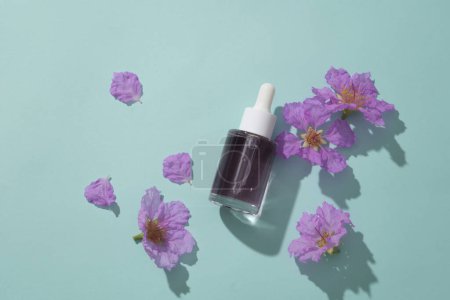 Glass bottle unlabeled with dropper cap, containing liquid and purple flowers decorated on a blue background. Mockup for cosmetic, serum of blueberry extract, moisturize and whiten skin naturally