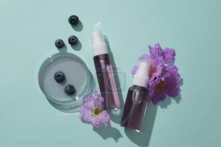 Promotional scene for cosmetic - an unlabeled bottle, essence in a petri dish and purple flower on a blue background. Blueberry extract have anti-aging and skin-regenerating effects.
