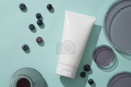 Top view of cosmetic bottle with blueberries and petri dishes filled purple liquid on blue background. Scene for advertising cosmetic from blueberry extract, rich in vitamin C and anthocyanins