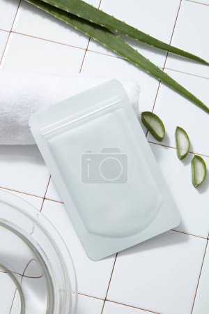 Photo for A facial sheet mask packaging mockup, white towel and fresh aloe vera leaves on white tile floor. Background for advertising cosmetic of aloe vera extract with bathroom concept. - Royalty Free Image