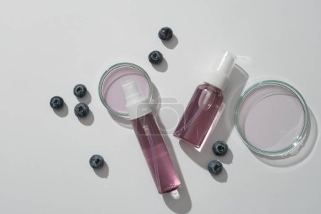 Two spray bottles unlabeled filled purple liquid, blueberries and petri dish on white background. Blueberries are rich in vitamin C and anthocyanins, which promote collagen production in the body.