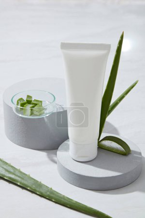 A white plastic tube unlabeled on round podium, fresh aloe vera on white background. Mockup scene for cosmetic of aloe vera extract - contains many nutrients such as antioxidants, vitamins (A, C, E)