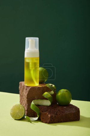 Front view of yellow spray bottle unlabeled placed on brown stone podiums and fresh limes decorated on dark background. Mockup scene for cosmetic product of lime extract. Minimal concept