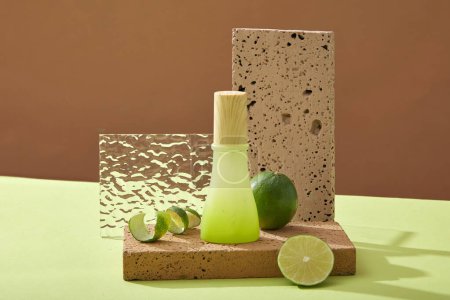 Front view of green glass bottle with wooden cap on brick podium, halves of lime and spiral lime peel on brown background. Mockup scene for product with lime ingredient. Space for design