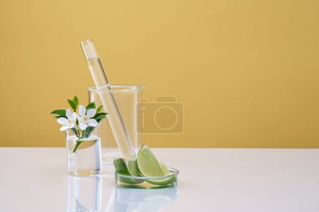 Lab glassware containing liquid, flower branch and lime slice on yellow background. Minimal empty display product. Lime extract contains a lot of vitamin C and nutrients for effective skin care.