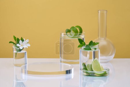 Minimal empty display product scene with lime. Laboratory glassware containing colorless liquid, flower, leaves and lime slices on yellow background. Empty space on transparent podium, front view.