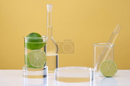 Transparent podium for display cosmetic product, lab glassware containing colorless liquid and fresh limes decorated on yellow background. Laboratory equipment and research natural extracts from lime