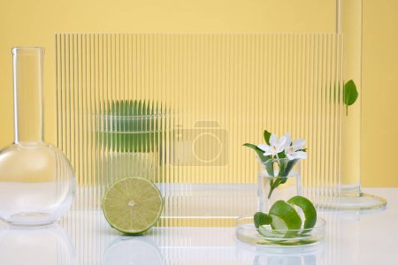 Téléchargez les photos : Scène publicitaire cosmétique d'extrait de citron vert avec podium transparent, verrerie de laboratoire, branche de fleur blanche et moitiés de citron vert décorées sur fond jaune. Espace pour le produit d'affichage. - en image libre de droit