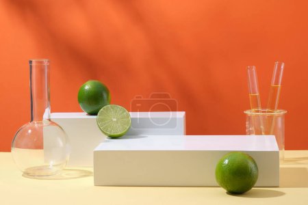 Front view of white rectangle cylinder podium, lab glassware containing yellow liquid and fresh limes decorated on orange background. Minimal empty display product presentation scene.