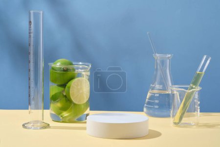 Front view of round empty podium, fresh limes and laboratory glassware containing transparent liquid on blue background. Blank space for display cosmetic of lime extract with minimal concept.