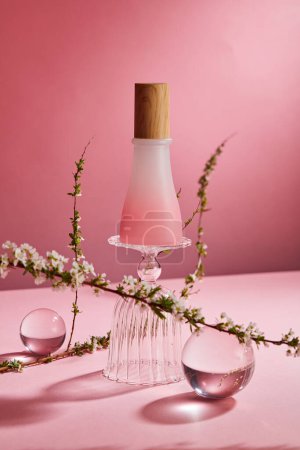 Szene-Attrappe für Kosmetikprodukte mit leerer Flasche ohne Etikett auf Glasbecher, transparenten Kugeln und Blumenzweigen auf rosa Hintergrund. Frontansicht, minimales Konzept, Beauty-Produktdesign.