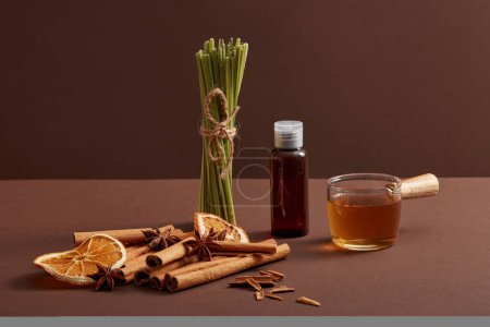 Brown bottle without label, lemongrass, cinnamon sticks, dried orange slice and anise on dark background. Mockup scene for product of natural essential oils extract. Healthy herb, natural flavoring