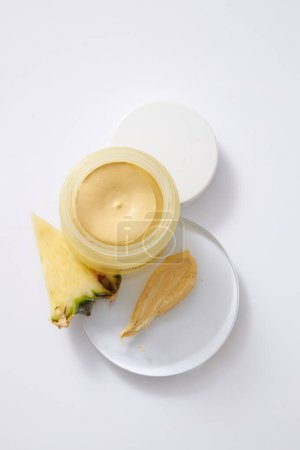 Photo for Top view of cream jar unlabeled, yellow cream texture on transparent podium and fresh pineapple slice decorated on white background. Cosmetic product mockup extracted from pineapple. Copy space - Royalty Free Image