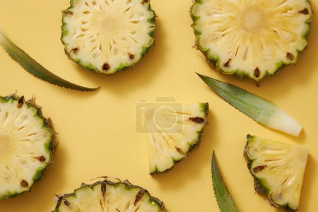 Tranches, cercles et triangles d'ananas (Ananas comosus) frais, feuilles vertes décorées sur fond jaune. Photo publicitaire, l'ananas est riche en vitamine C apporte des bienfaits inattendus à la peau.