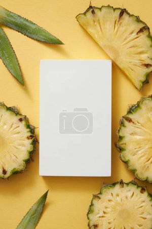 Photo for A white rectangle empty podium surrounded by fresh pineapple (Ananas comosus) slices and green leaves on yellow background. Empty space for cosmetic of pineapple extract presentation. - Royalty Free Image