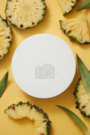 Vue de dessus du podium rond blanc entouré de tranches d'ananas frais (Ananas comosus) et de feuilles vertes sur fond jaune. Piédestal pour produit cosmétique avec des ingrédients de l'affichage de l'ananas.