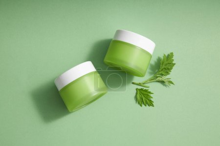 Top view of two green cream jar unlabeled mockup for product of moisturizers, masks or scrubs. Fresh wormwood leaves on green background. Mockup scene for cosmetic of wormwood extract.