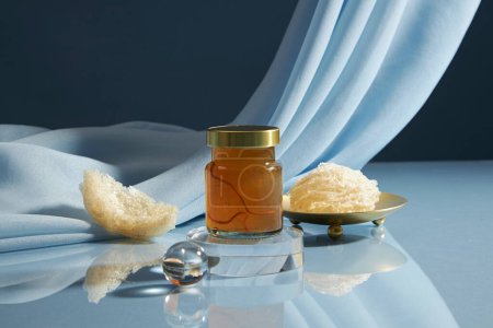 A glass bottle unlabeled containing bird nest water on transparent podium, some raw edible bird nest decorated on blue soft fabric background. healthcare food, front view, copy space