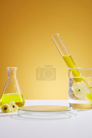 Front view of laboratory equipment filled with yellow liquid in erlenmeyer flask and test tube, white flower in beaker and transparent empty podium on yellow background. Space for display product.
