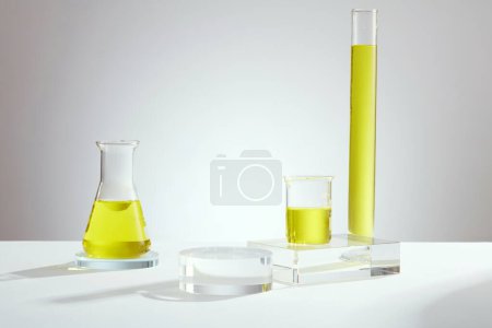 Laboratory concept with erlenmeyer flask, beaker and test tube containing yellow liquid and empty transparent podium on light background. Background for the presentation of cosmetic products