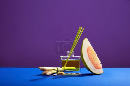Advertising backdrop for products and cosmetics with ingredients from lemongrass and pink pomelo. Front view of fresh lemongrass, glass bottle of essential oil and pomelo pieces on purple background.