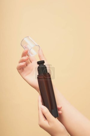 Photo for Scene of a female hand holding an amber plastic bottle without label, the other hand opening the mouth of the bottle on beige background. Mockup for cosmetic product, advertising photo - Royalty Free Image